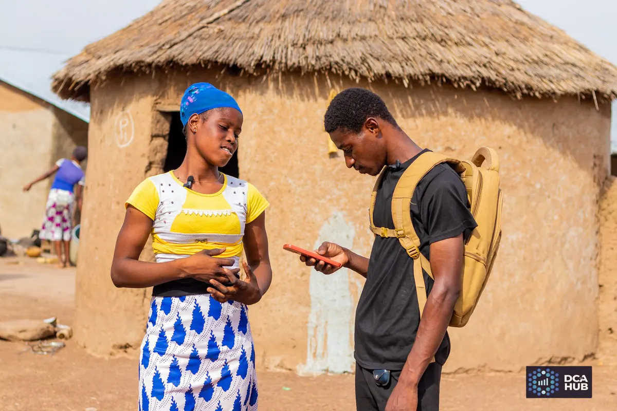 An enumerator shows a respondent something on a smartphone during digital data collection in a rural compound in Gushegu